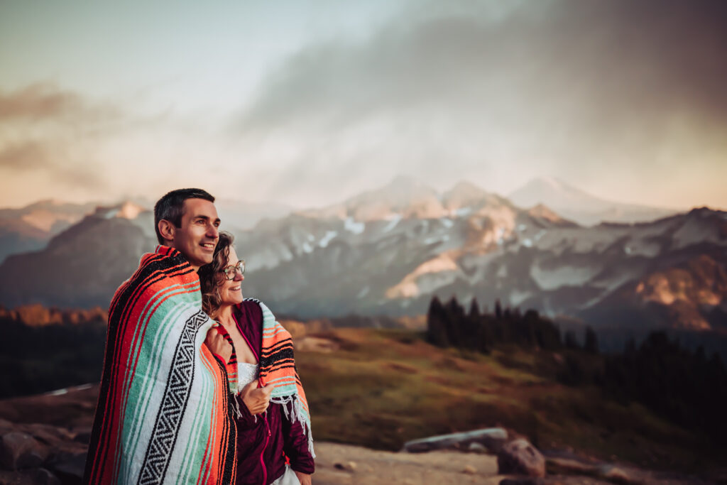 A wedding couple wrapped in a blanket watching the sunset in the mountains