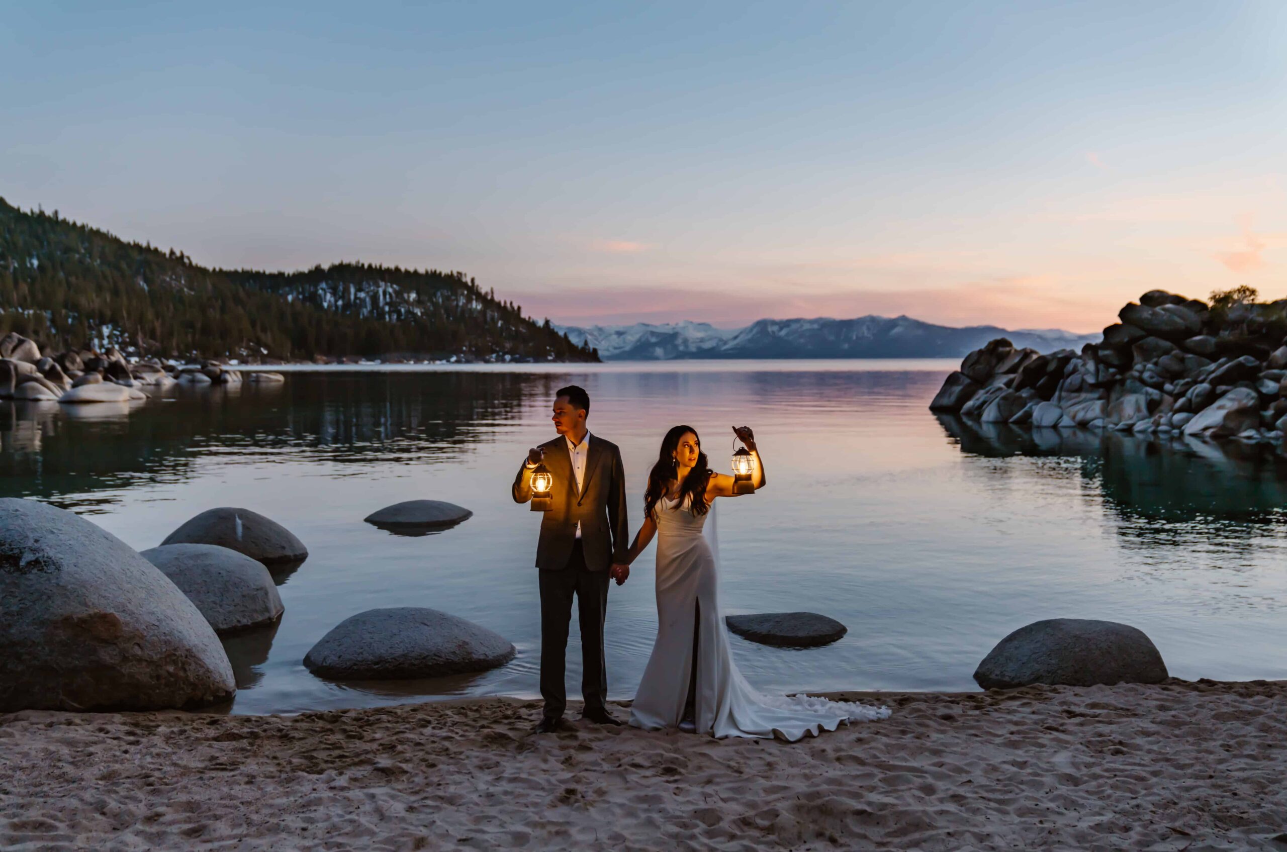 A bride and groom standing on the beach in Lake Tahoe holding lanterns in the dim light for their elopement in Tahoe