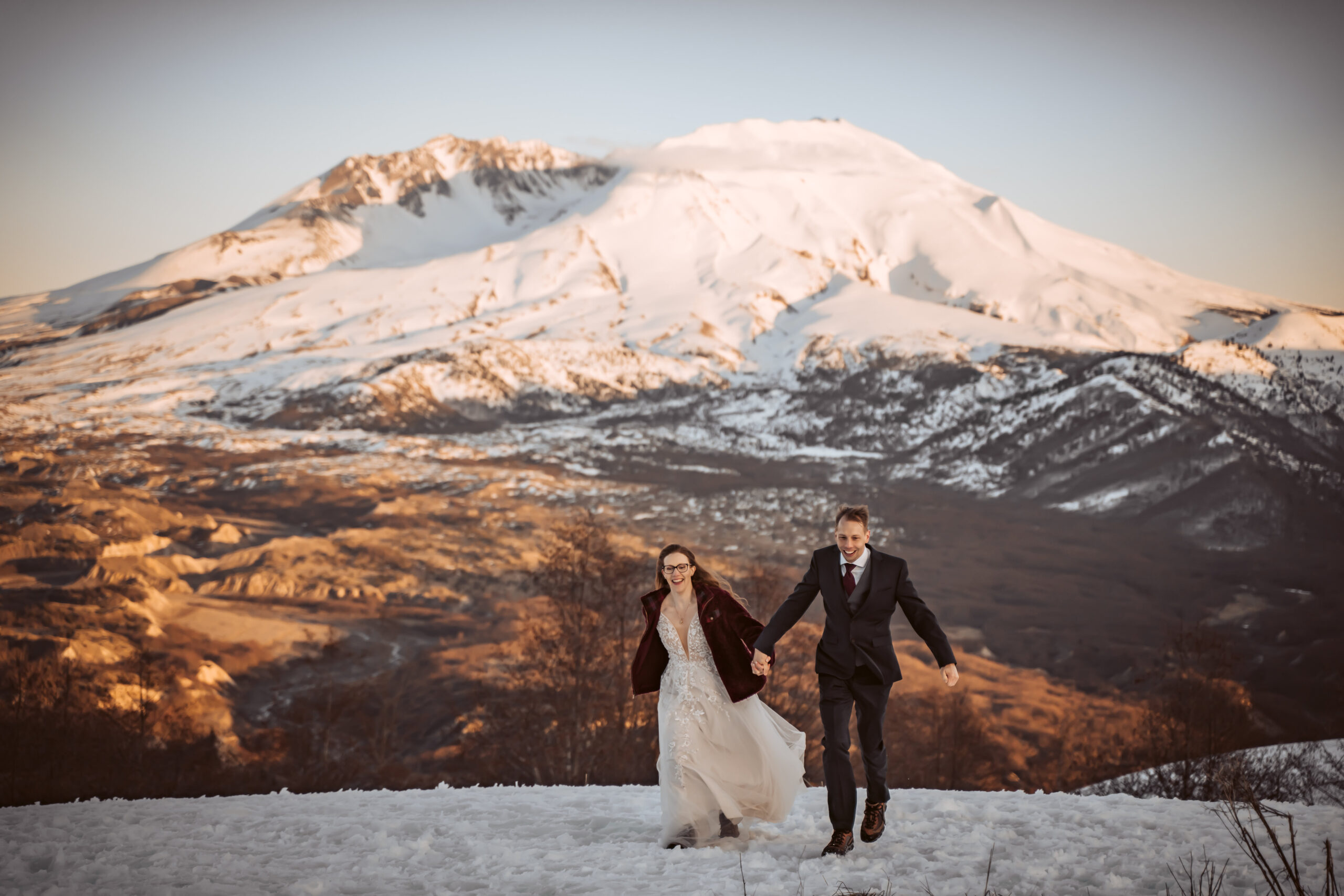 a couple running in the snow and holding hands with Mount Saint Helen's in the backdrop for their elopement day
