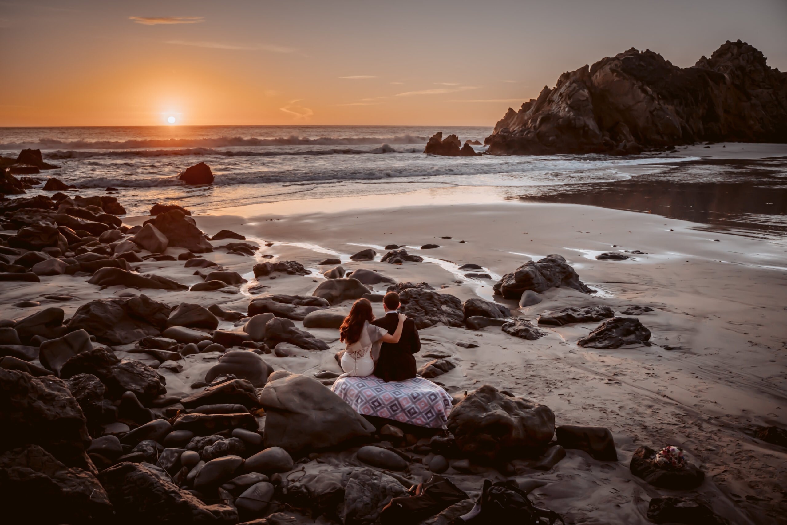 A couple sitting on the rocks looking out onto the sunset on the beach in California for their elopement