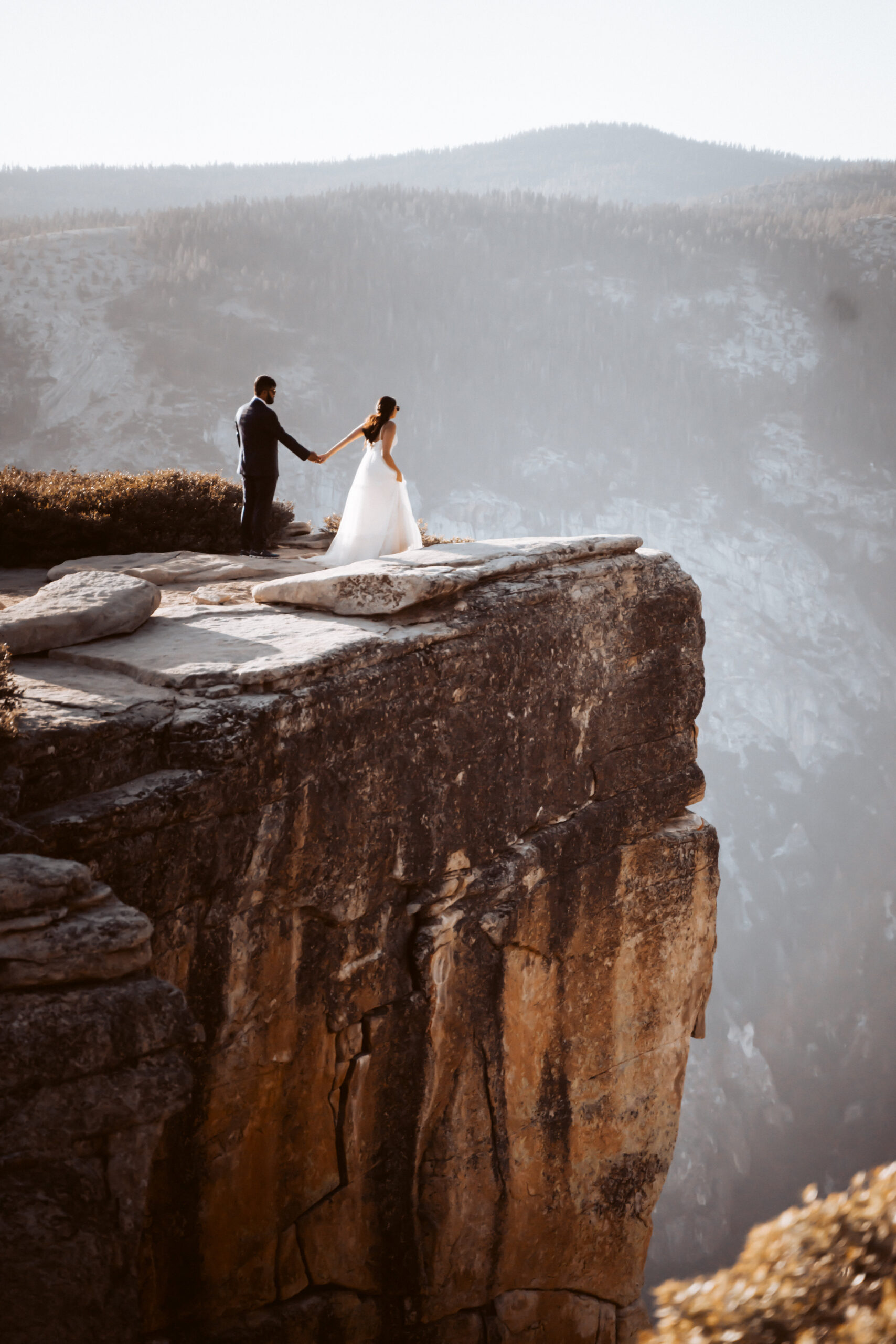A bride and groom standing on a large rock ledge overlooking Yosemite Valley for their Elopement day 