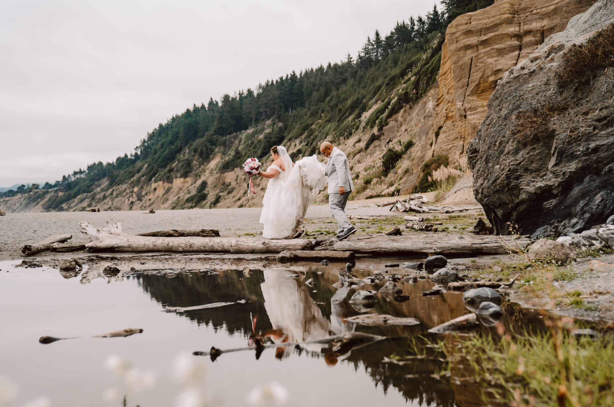 A bride and groom walking over a log to get the beach for their elopement ceremony on the California coast