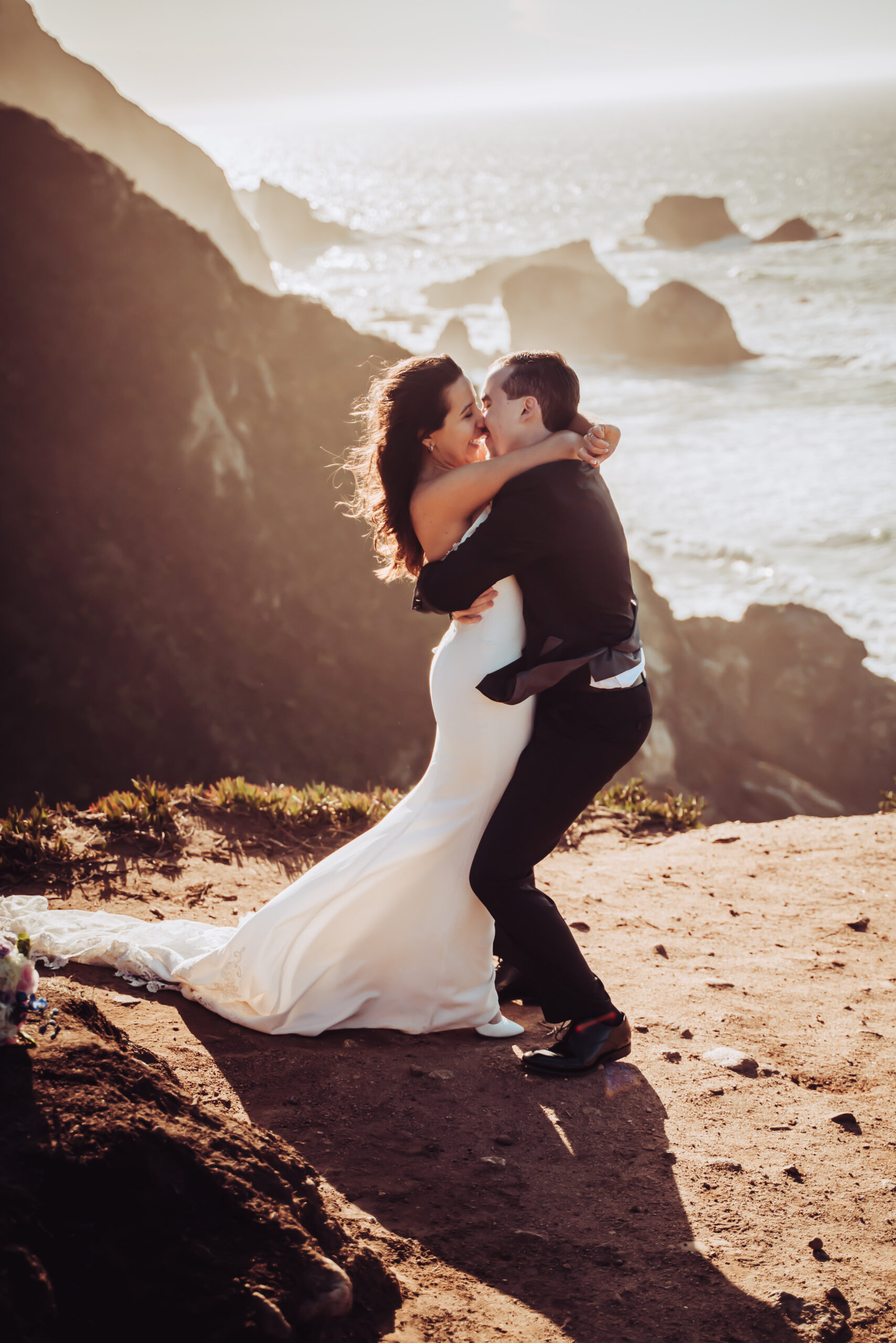 A groom holding his bride overlooking the cliffs and sunset of Big Sur on their Elopement day