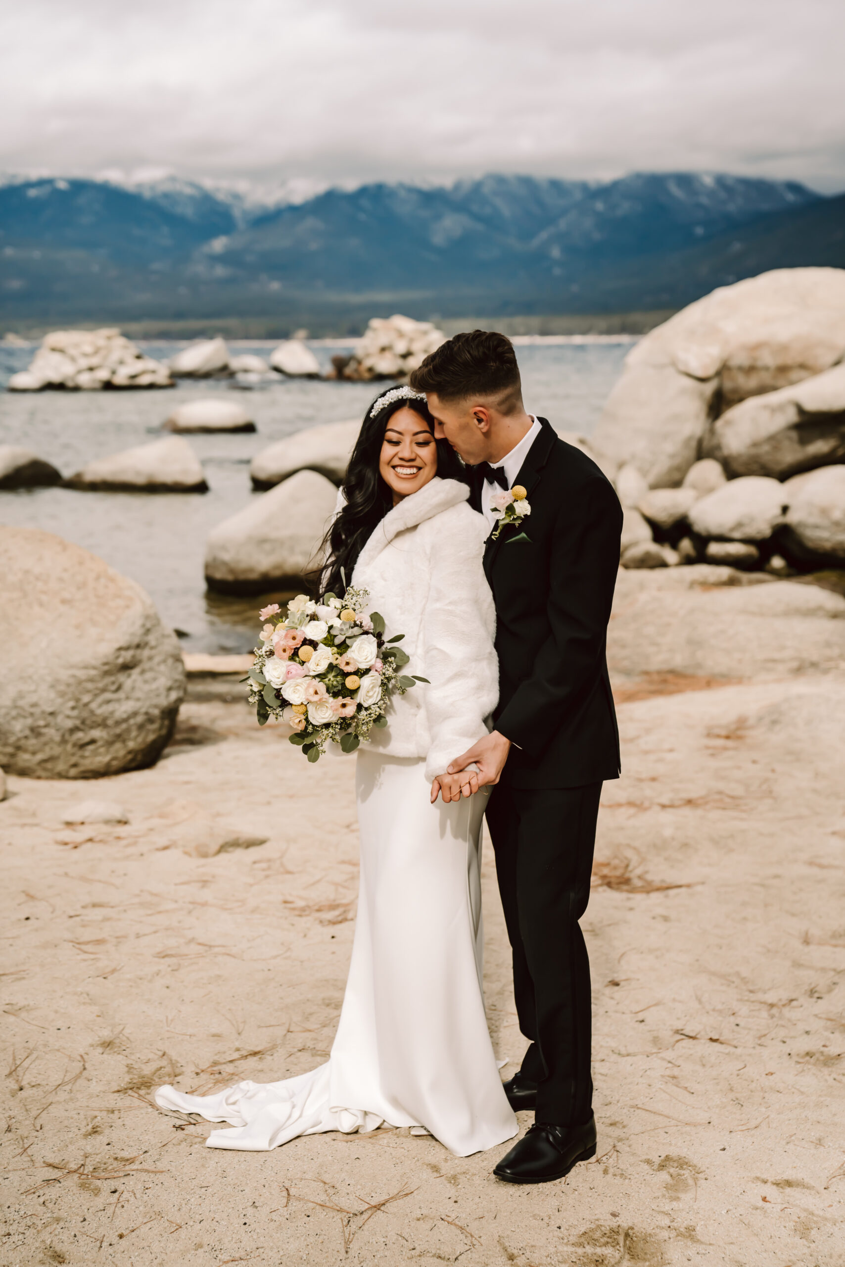 A groom kissing his bride on the beach of Sand Harbor with the mountains in the backdrop for a winter Elopement in Lake Tahoe