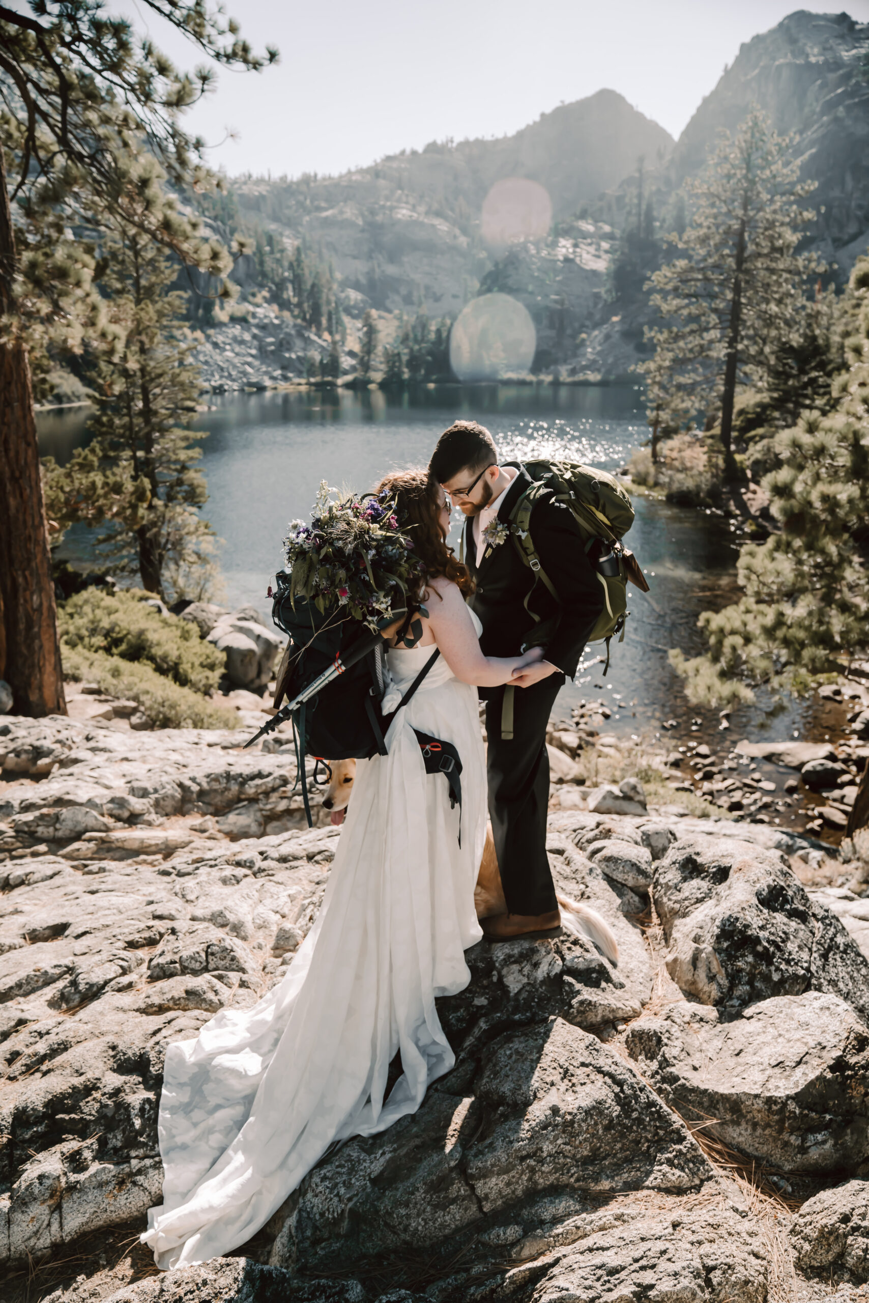 a bride and groom with hiking backpacks kissing in front of a mountain lake backdrop for their elopement day