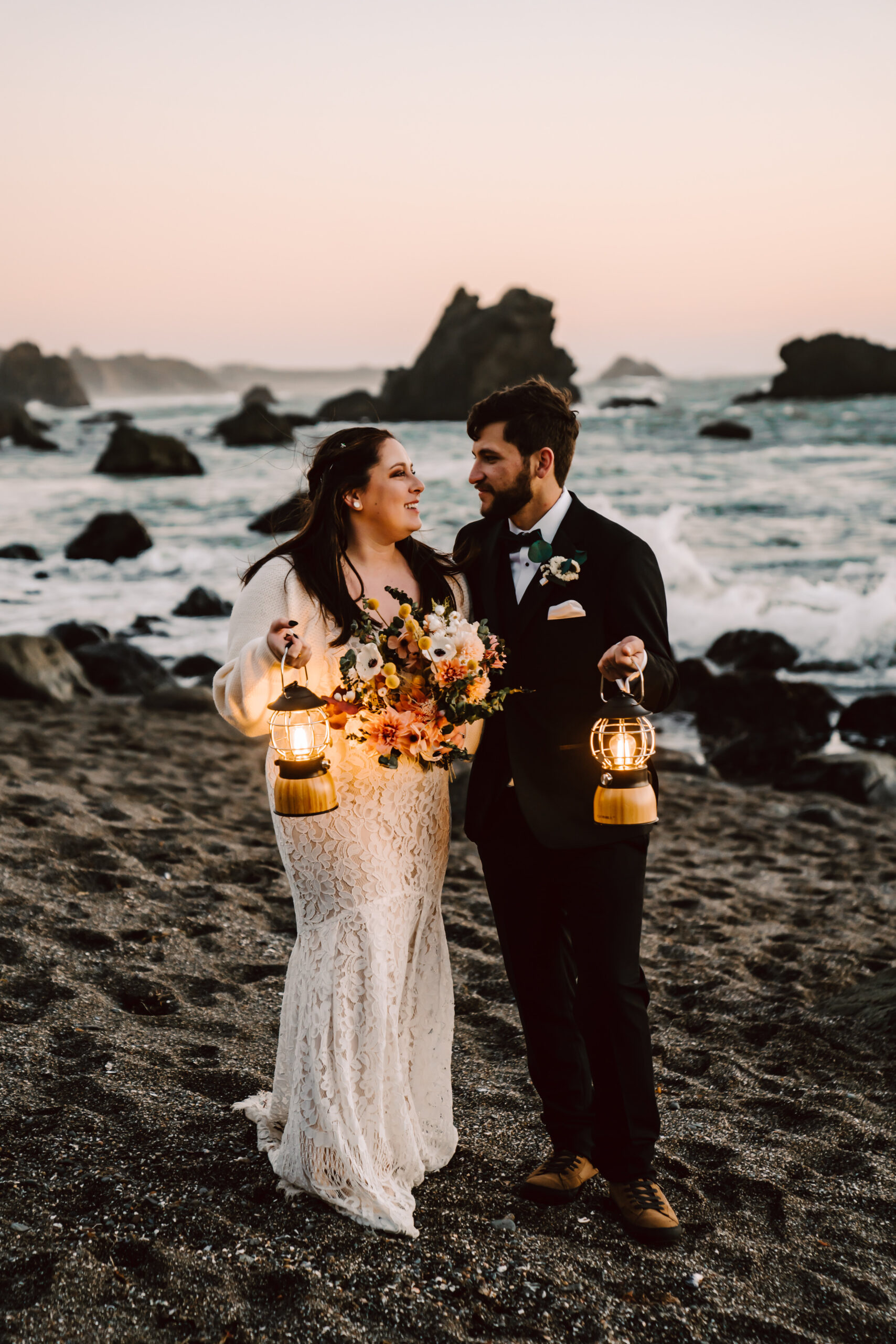 A bride and groom holding lanterns walking on the beach at sunset during their elopement day