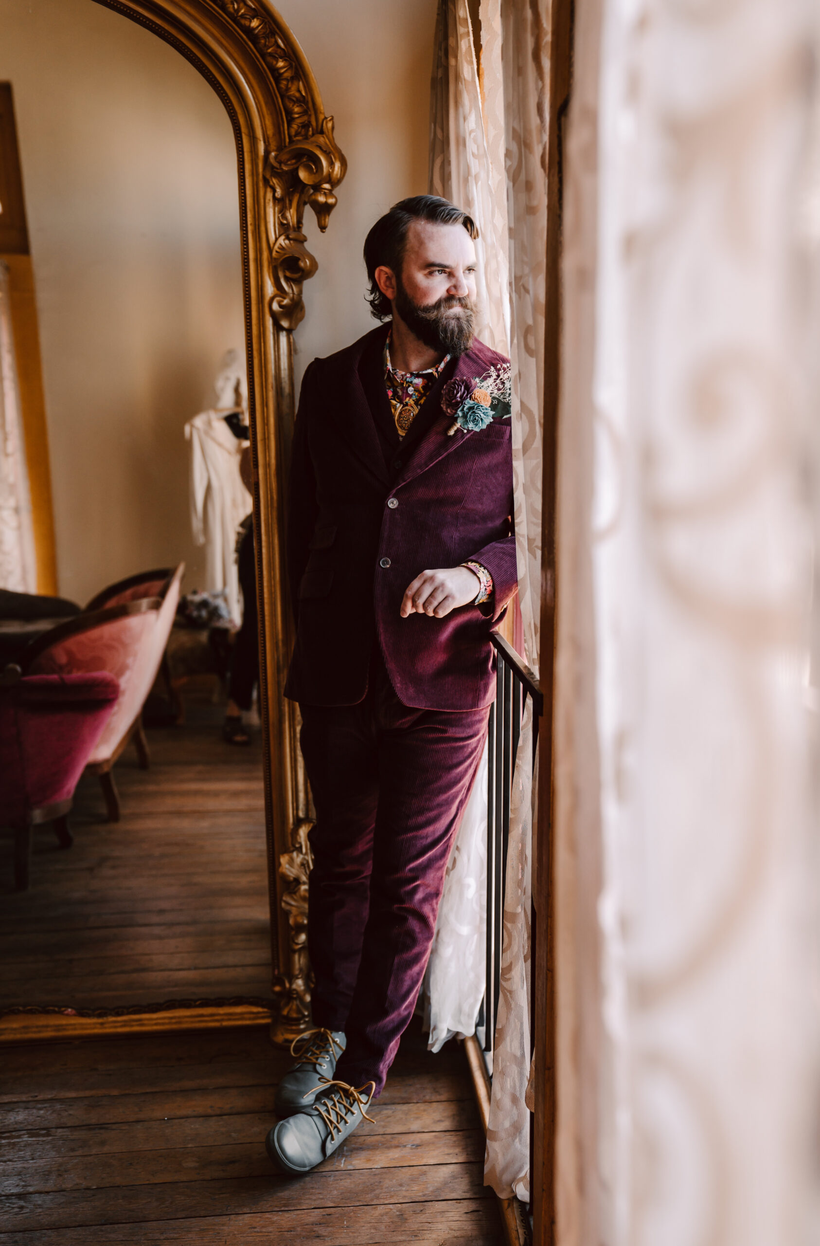 A groom wearing a deep red corduroy tux with floral tie looking out the window