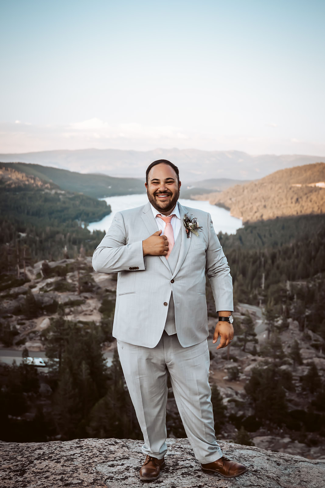 A groom wearing a light grey elopement suit with the mountains and the lake behind him