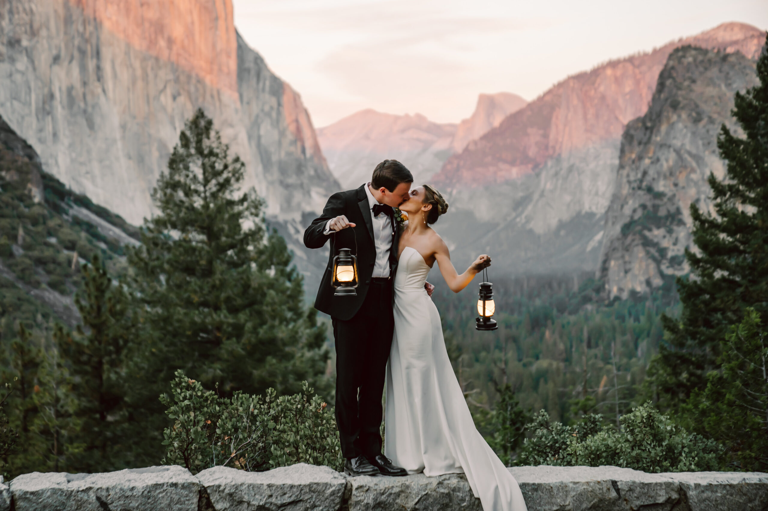 A bride and groom kissing and holding lanterns in front of the sunset at Yosemite Valley for their elopement day