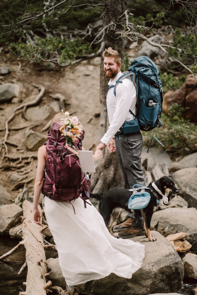An adventure elopement wedding couple with hiking backpacks helping each other cross a stream while their dog watches