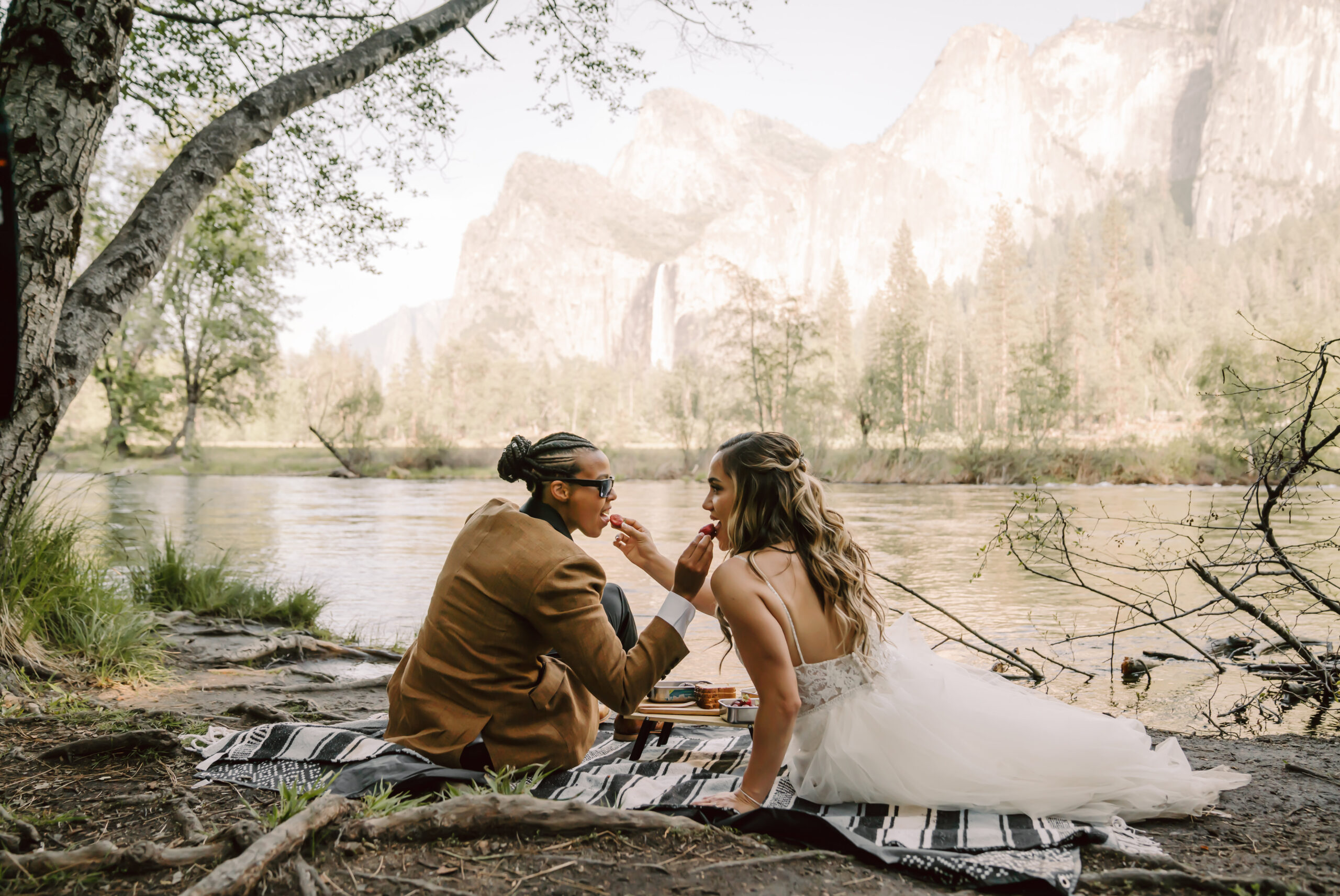 Two brides sitting on a mountain blanket next to a river in Yosemite Valley having a picnic for their elopement day and feeding each other