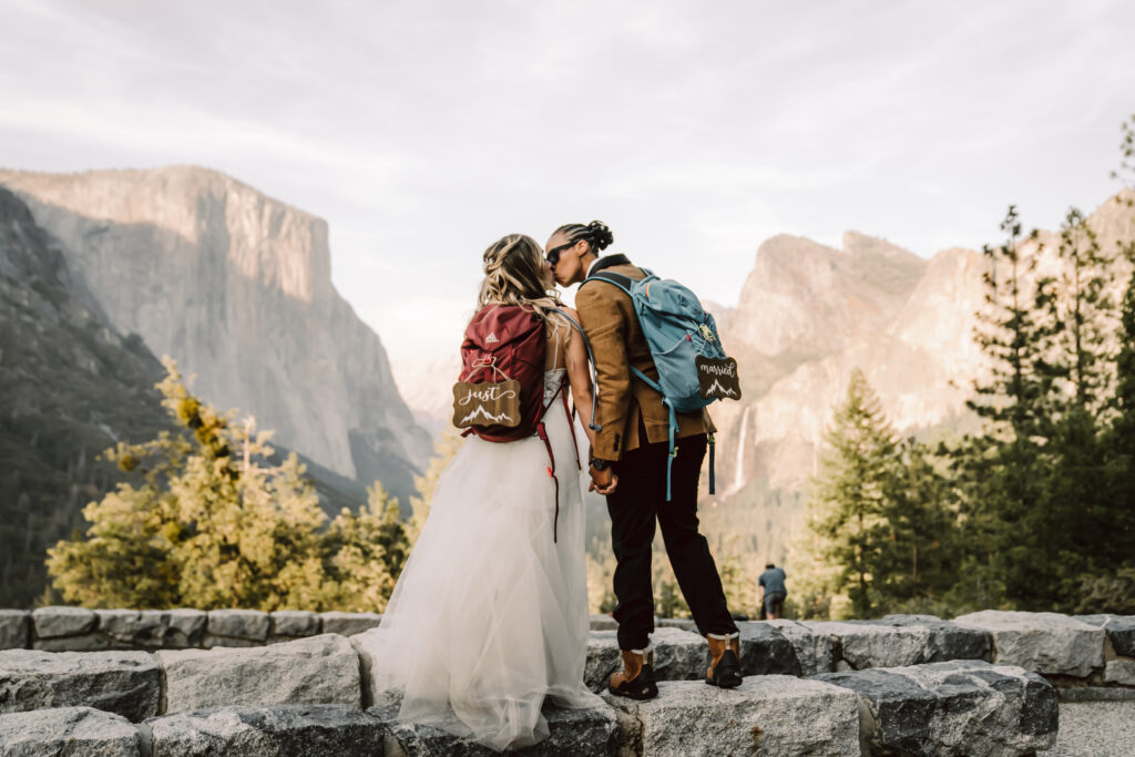 two brides wearing hiking backpacks and kissing overlooking Tunnel View in Yosemite for their Elopement day
