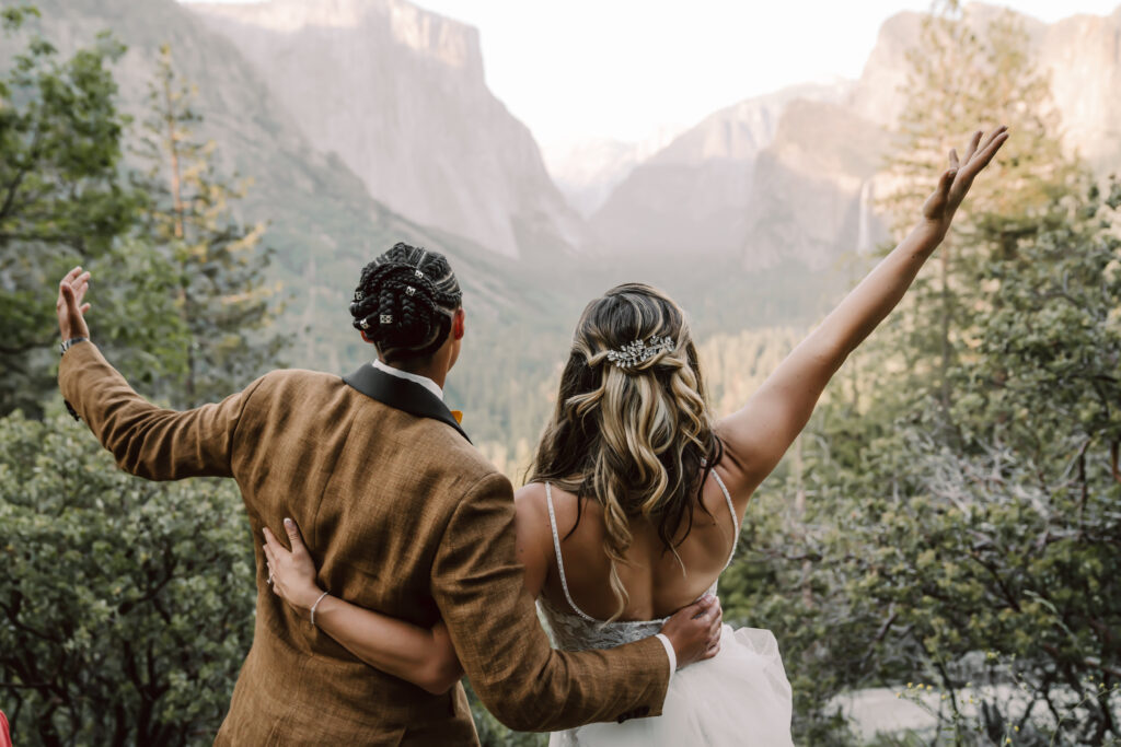 Two brides looking out onto Tunnel View for Yosemite Valley with their arms in the air celebrating their elopement day