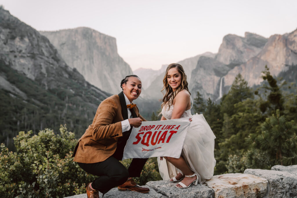 two brides squatting in front of tunnel view holding a sign that says drop it like a squat for their Yosemite Elopement day  