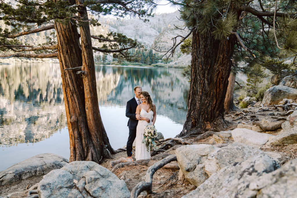A bride and groom standing in between two trees with a big reflection on eagle lake in Lake Tahoe behind them