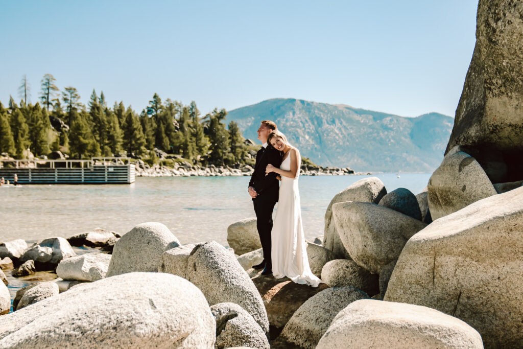 A Bride and groom standing on boulders in Lake Tahoe with a pier and a mountain in the backdrop