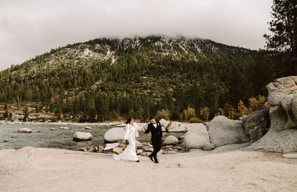 A wedding couple walking on the beach with a mountain backdrop with clouds on it