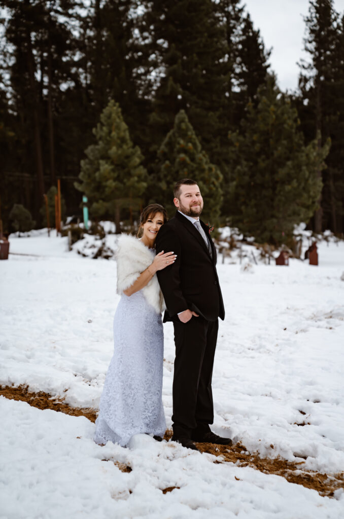A bride holding her groom from behind and standing in the snow with the forest in the backdrop