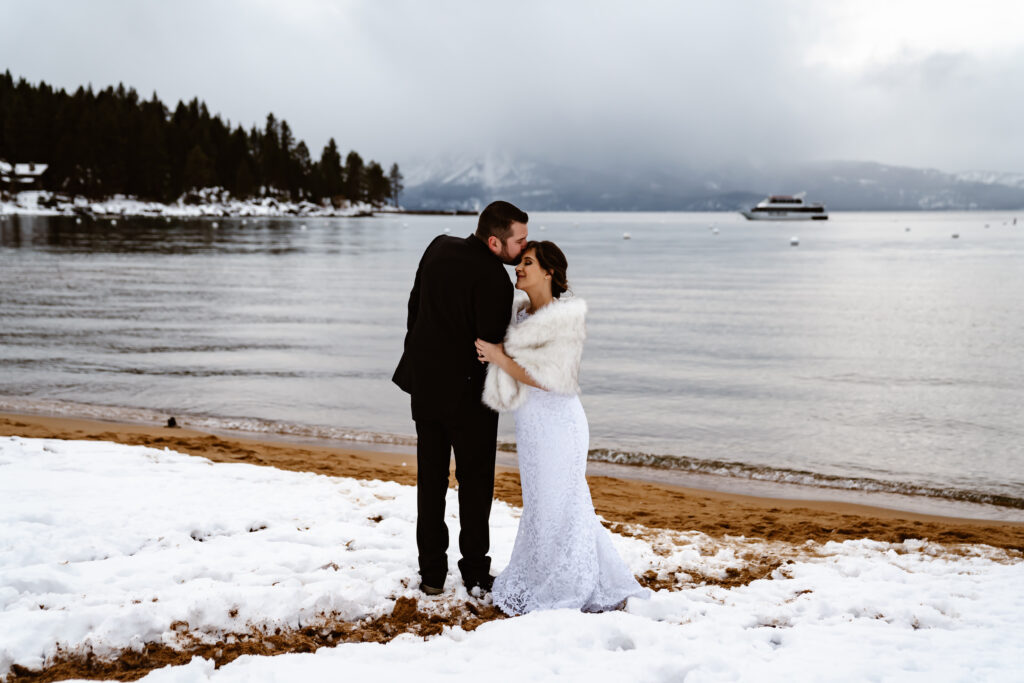 A groom kissing his bride on the head and standing in the snow with Lake Tahoe in the backdrop
