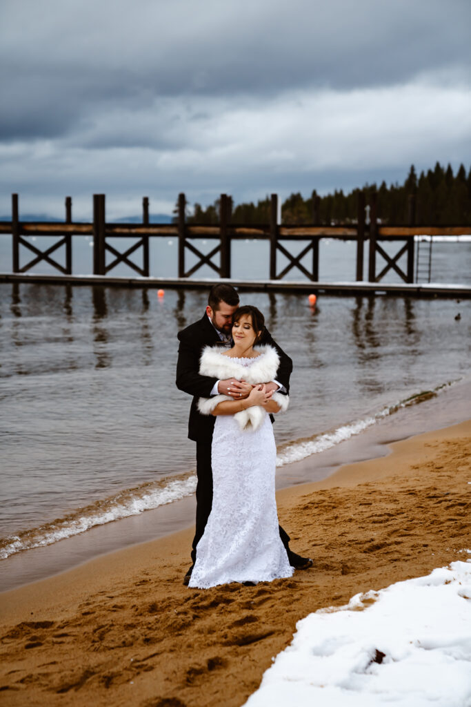 A groom holding his bride from behind and standing in front fo the dock at Zephyr Cove in Lake Tahoe for their wedding day