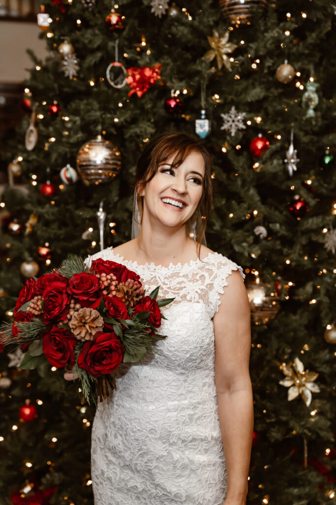 A bride standing next to a Christmas Tree holding a bouquet of red flowers