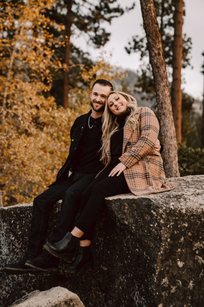 an engaged couple sitting on a boulder with their heads together smiling