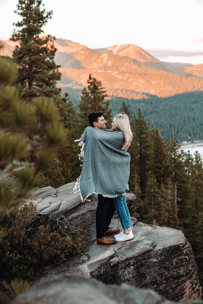 an enaged couple  wrapped in a blanket standing on some boulder overlooking the mountains during blue hour alpenglow