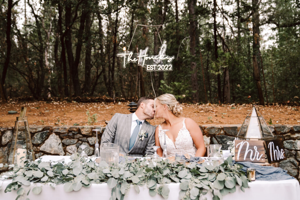 bride and groom kissing at the head table under a neon sign that says their new last name at their wedding reception at the Roth Estate in Nevada City