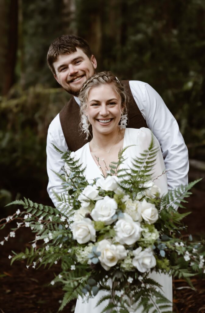 a wedding couple in the rainforest smiling at the camera and holding a fern bouquet 