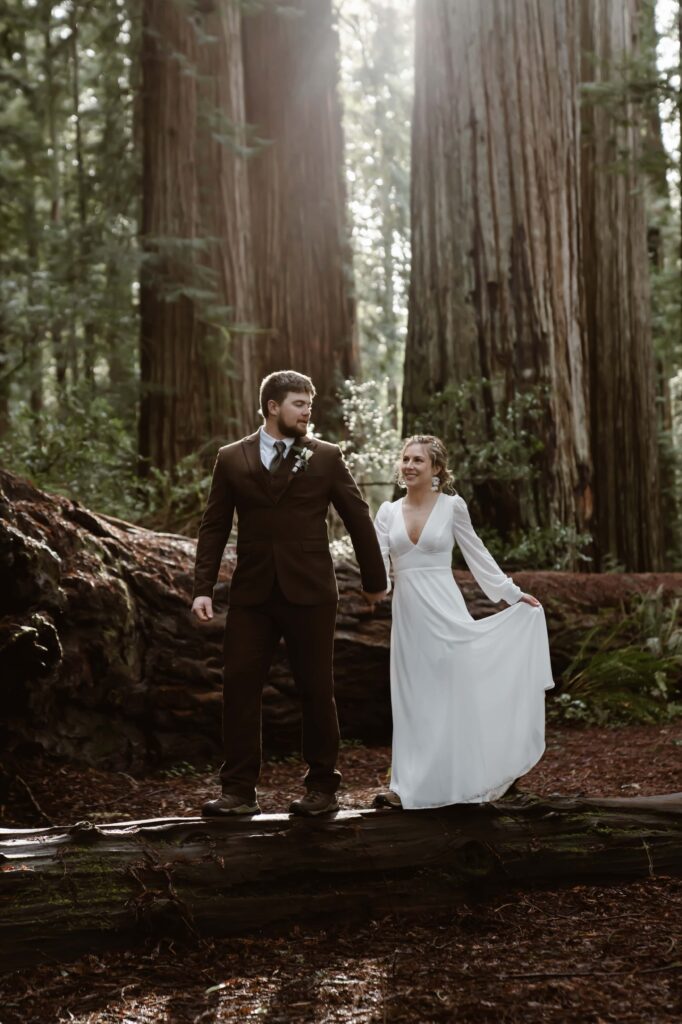  a bride and groom walking across a log in front of the giant redwoods on their elopement day