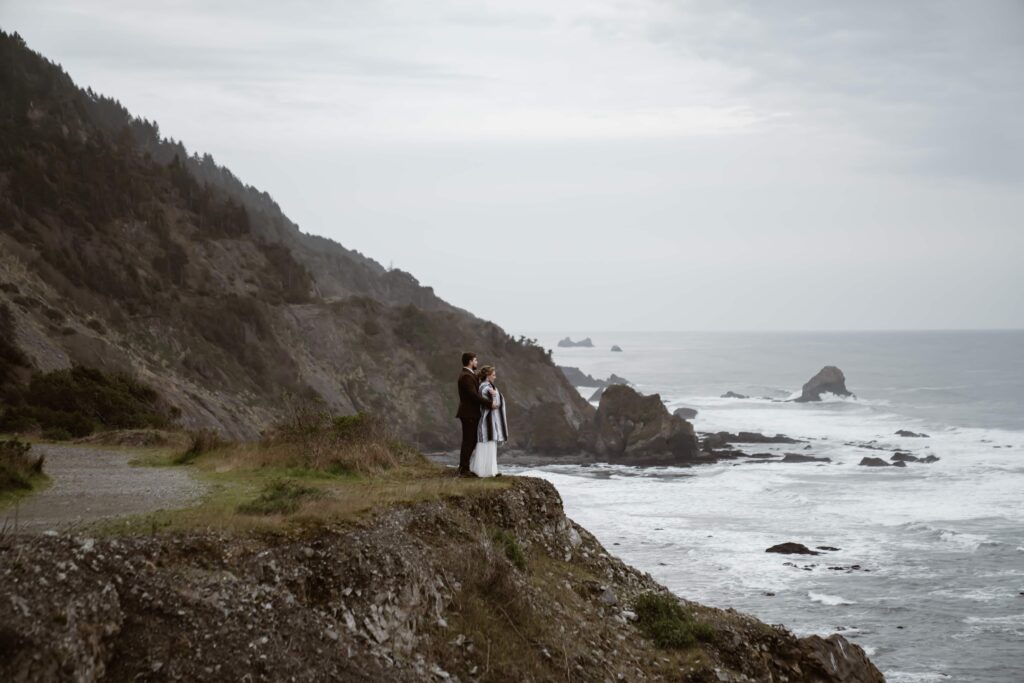 a bride wrapped in a blanket standing on a cliff overlooking the ocean at Enderts Beach in Northern California