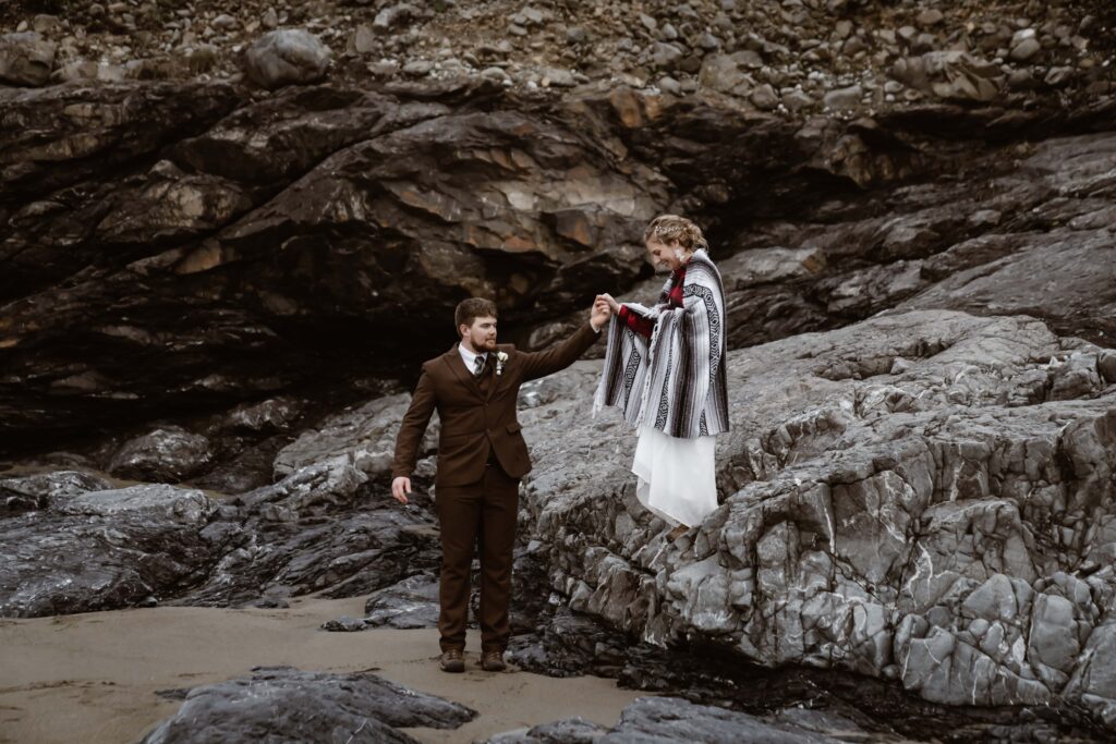 a groom helping his bride down the rocks at Enderts Beach for their elopement day