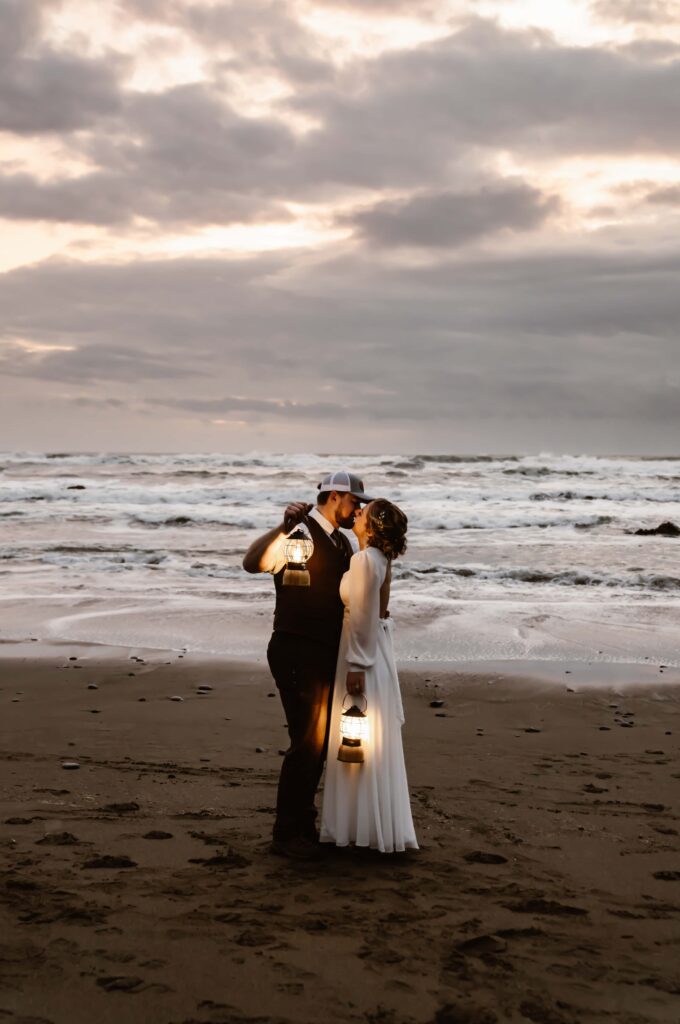 an elopement couple standing on the beach at sunset holding lanterns and kissing