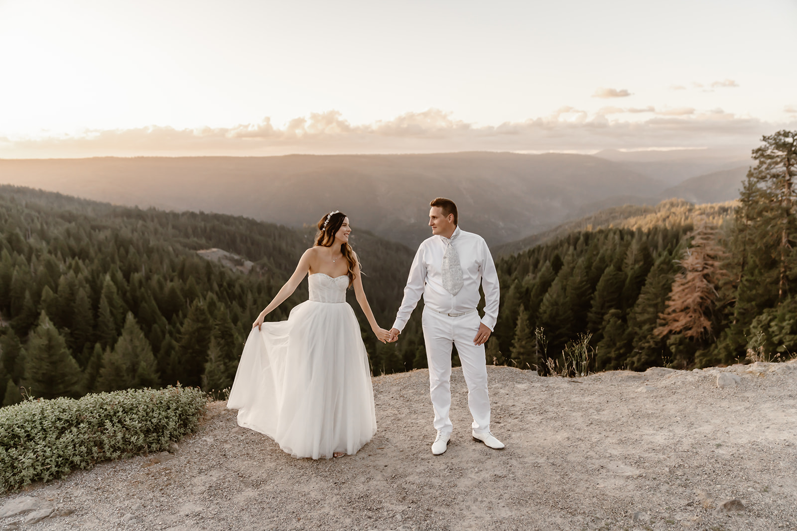 Bride and groom hold hands while smiling at each other with mountain backdrop