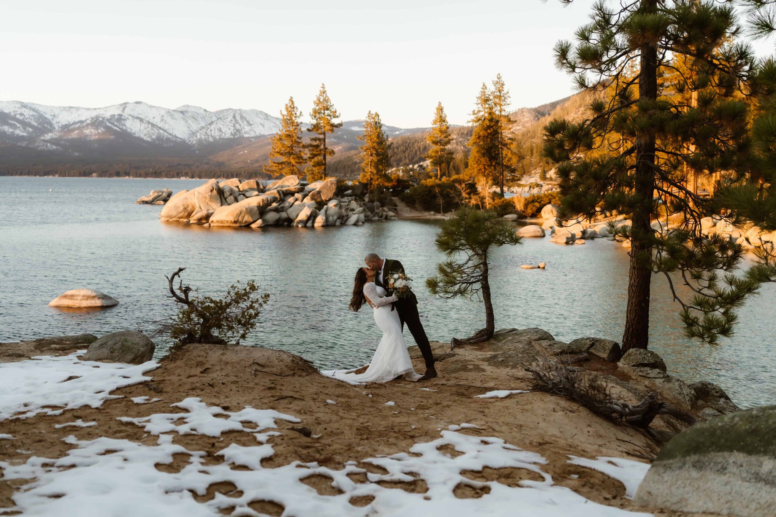 Lake Tahoe elopement couple kissing at Sand Harbor with snowy mountains, pine trees, and granite boulders along the shoreline.